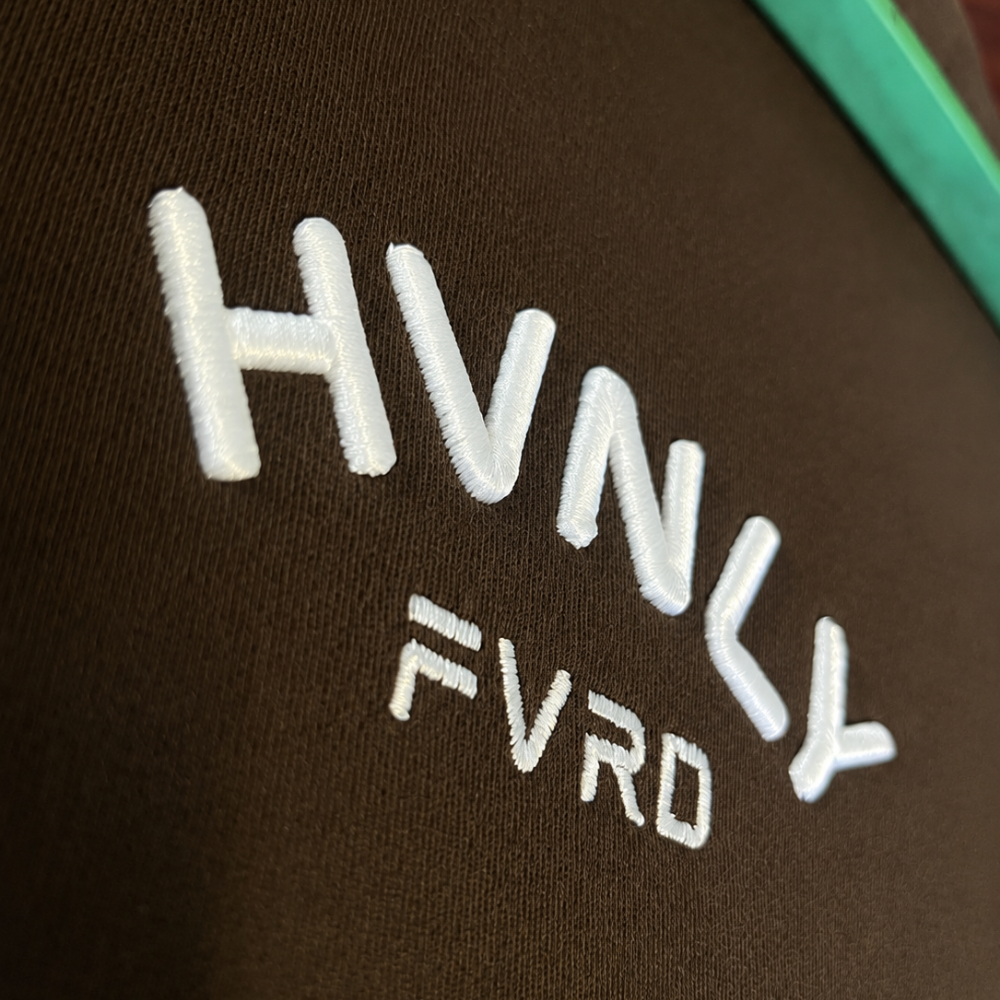 Close-up of white HVNLY FVRD lettering embroidered on a brown hoodie