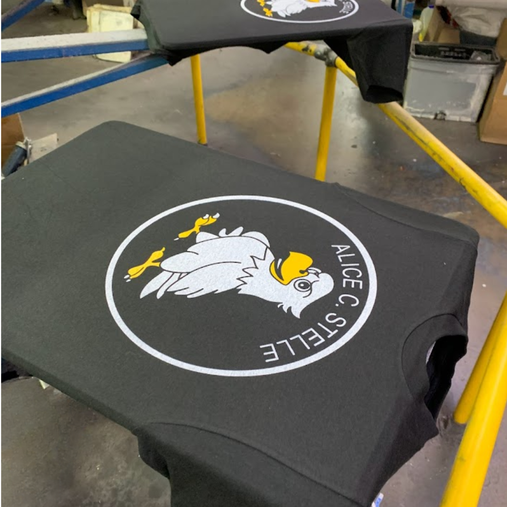 Black screen printed shirts on the press with a white and yellow mascot graphic